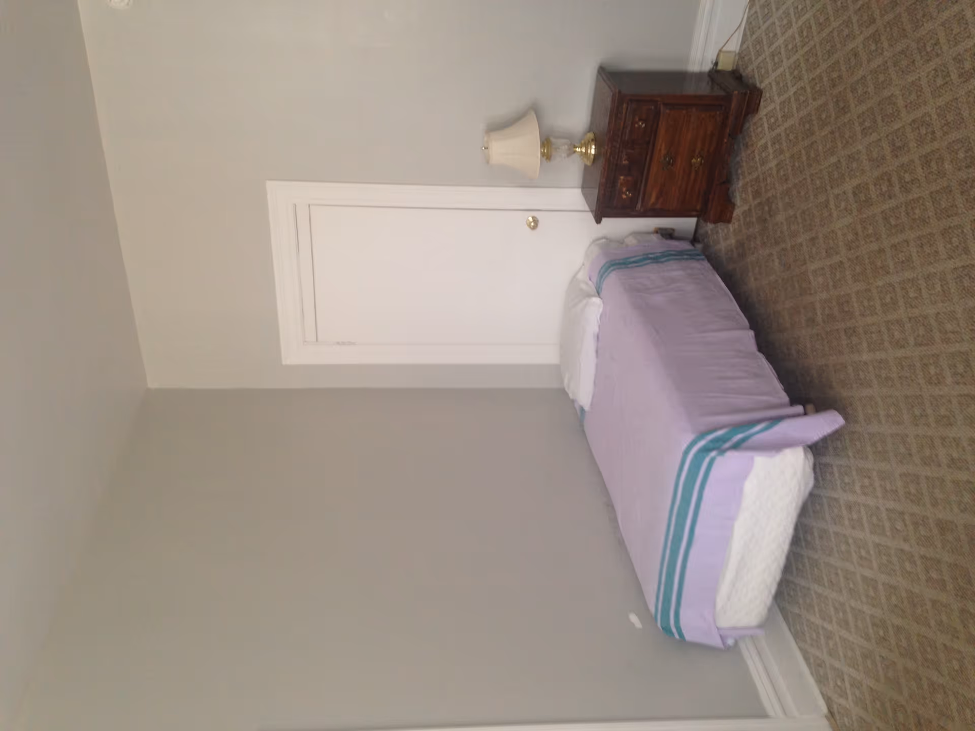 A small bedroom with a single bed covered with a light purple blanket with teal stripes, a white pillow, a wooden nightstand with drawers, and a lamp with a white shade. The walls are painted light gray and there is a white door behind the bed. The floor is carpeted with a patterned design.