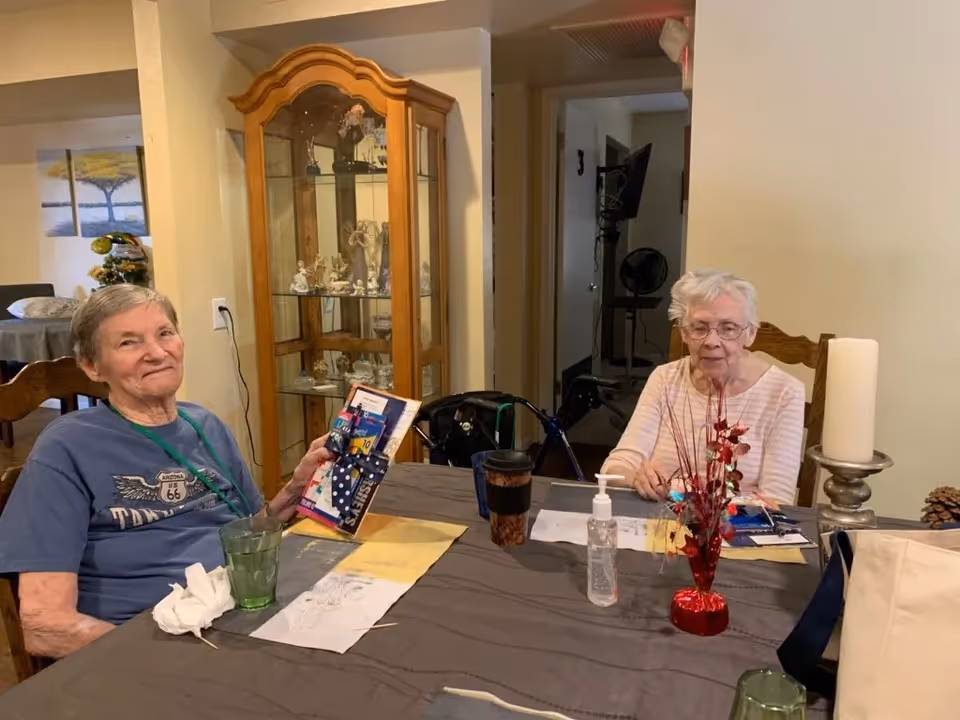 Two elderly women sitting at a dining table in a room with a wooden display cabinet filled with figurines behind them. One woman is holding a decorated card and smiling, while the other woman is looking down at the table. The table has a gray tablecloth, a red decorative centerpiece, a large candle on a holder, a bottle of hand sanitizer, and some cups.