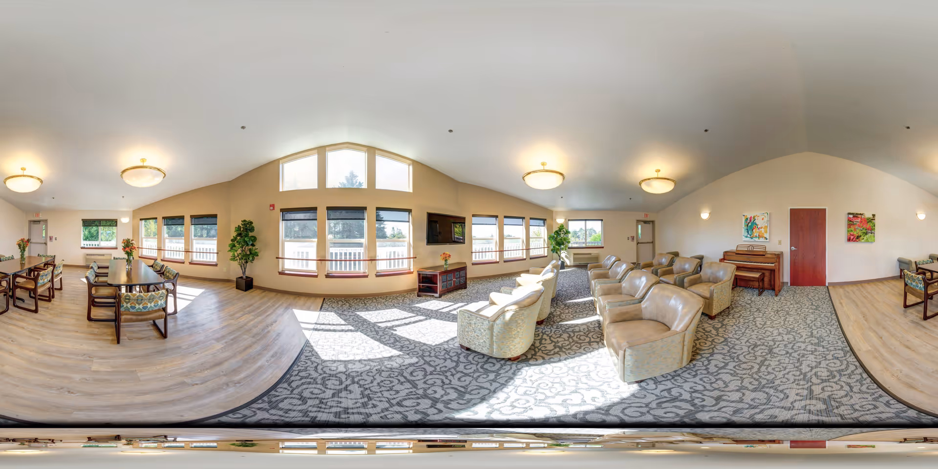 A spacious, well-lit common area in a senior living facility featuring a seating area with multiple beige armchairs arranged in rows on a patterned carpet. Large windows allow natural light to fill the room. There is a flat-screen TV mounted on the wall above a wooden cabinet, and a piano against the far wall. To the left, there are dining tables with chairs on a wood floor, decorated with flower vases. The walls are light-colored with some colorful artwork and potted plants placed near the windows.
