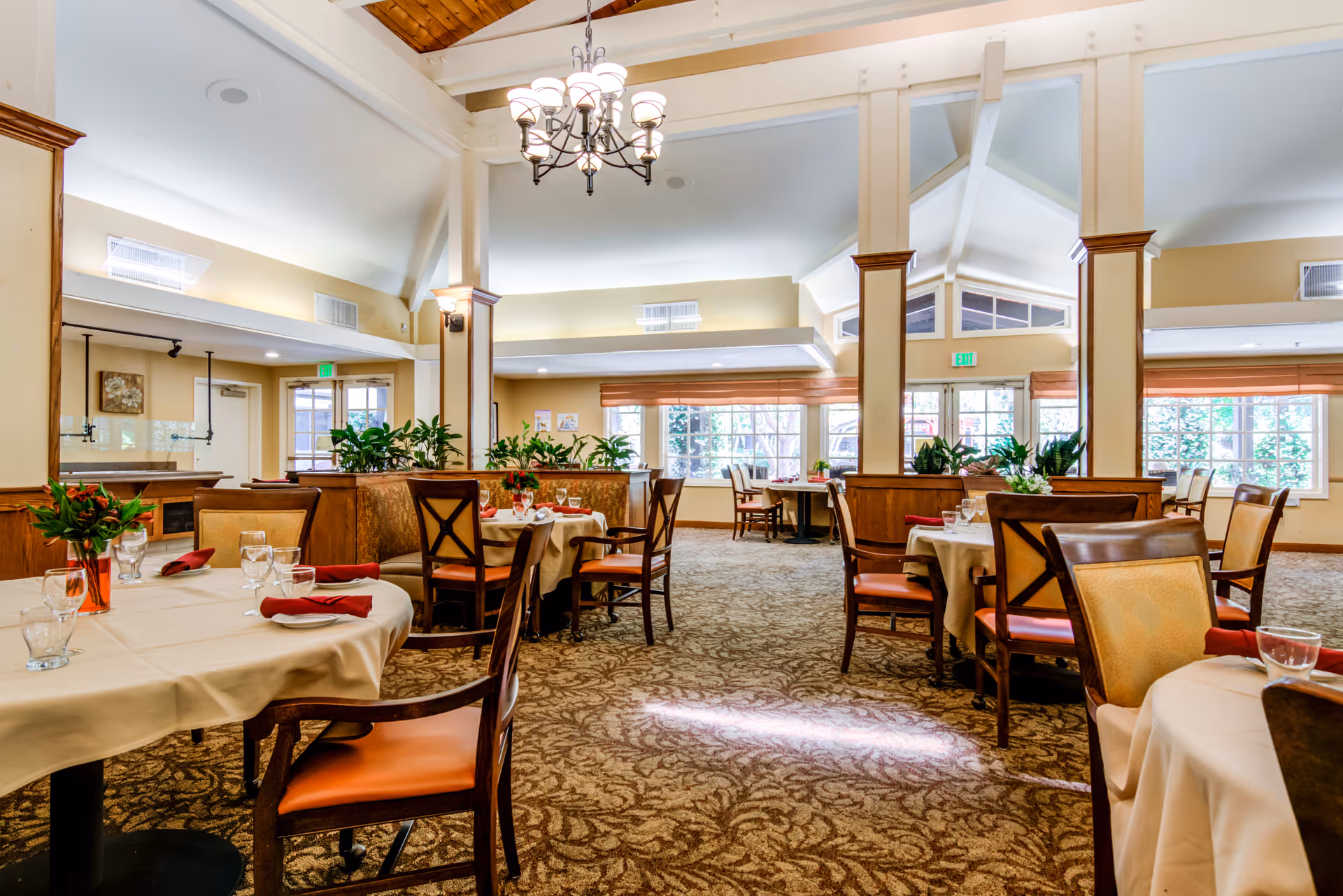 A spacious and well-lit dining room with multiple tables covered in beige tablecloths, each set with glasses, napkins, and small floral centerpieces. The room features high vaulted ceilings with wooden beams, large windows allowing natural light to fill the space, and a chandelier hanging from the ceiling. The carpet has a patterned design, and there are wooden chairs with cushioned seats around the tables.
