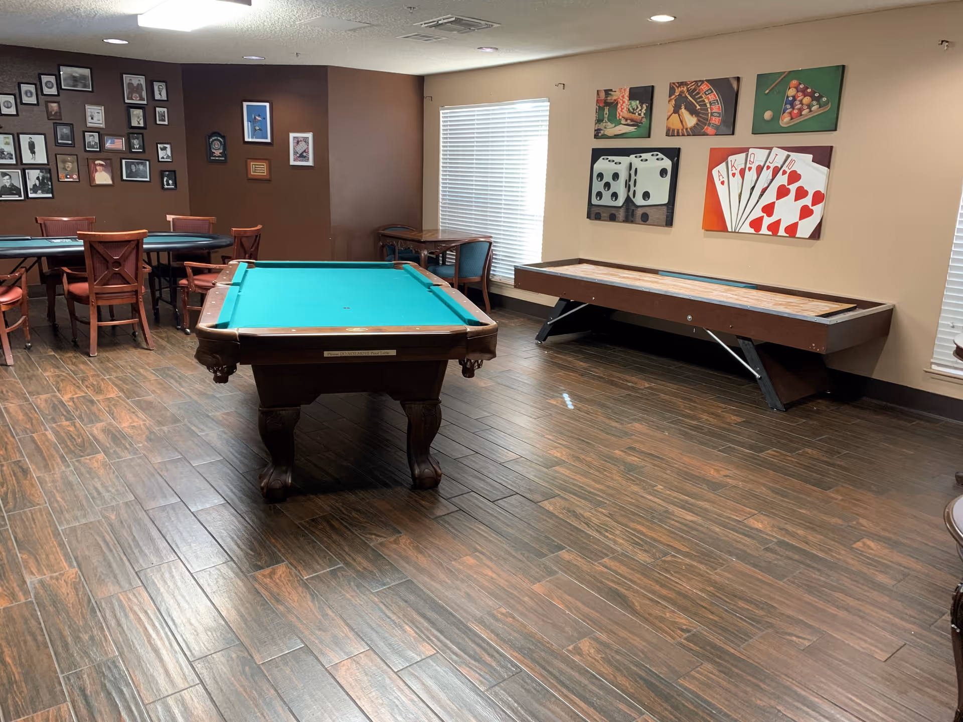 A recreational room with a pool table in the center, a shuffleboard table against the wall, and a card table with chairs in the background. The walls are decorated with framed pictures and artwork featuring dice, playing cards, and other game-related themes. The floor has a wood-like finish, and there are windows with blinds letting in natural light.