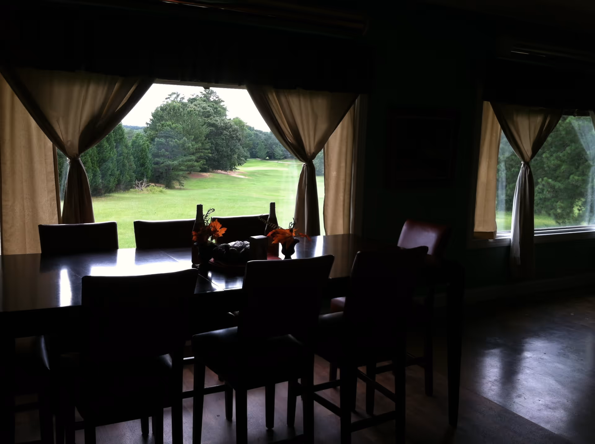 A dining room with a dark wooden table and several chairs. The table has decorative items including small vases with orange flowers. Large windows with beige curtains tied back reveal a green outdoor landscape with trees and grass.