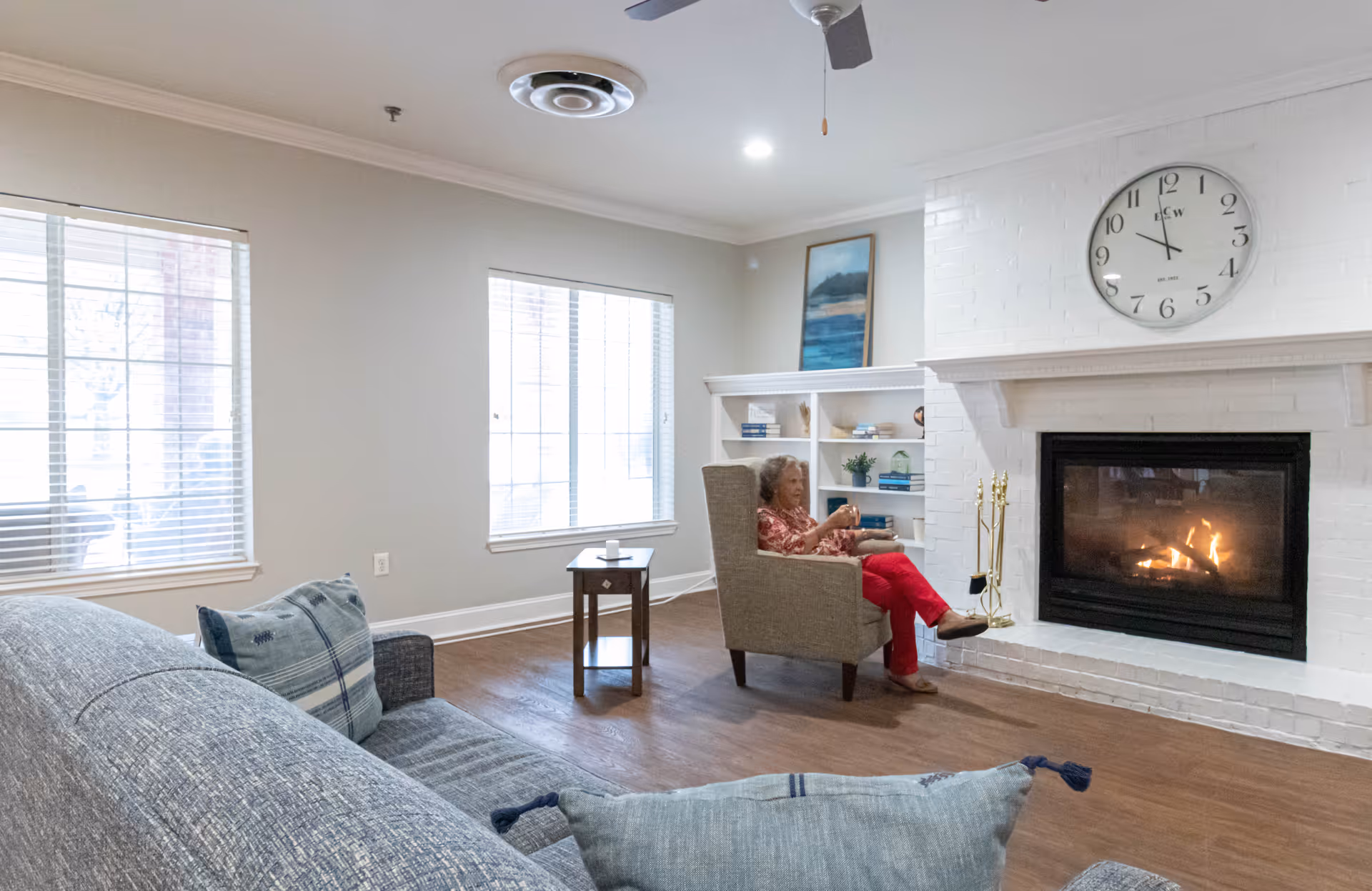 A cozy living room with a gray couch and pillows in the foreground, a woman sitting in a beige armchair near a white brick fireplace with a fire burning, a large clock above the fireplace, two large windows with blinds, a small wooden side table, and a white built-in bookshelf with decorative items and books.