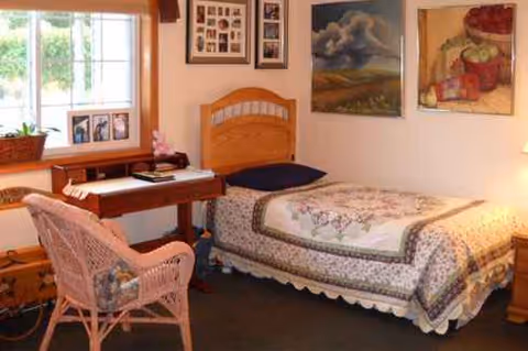 Cozy bedroom inside Cottagewood Senior Communities with a bed, wicker chair, desk, and artwork.