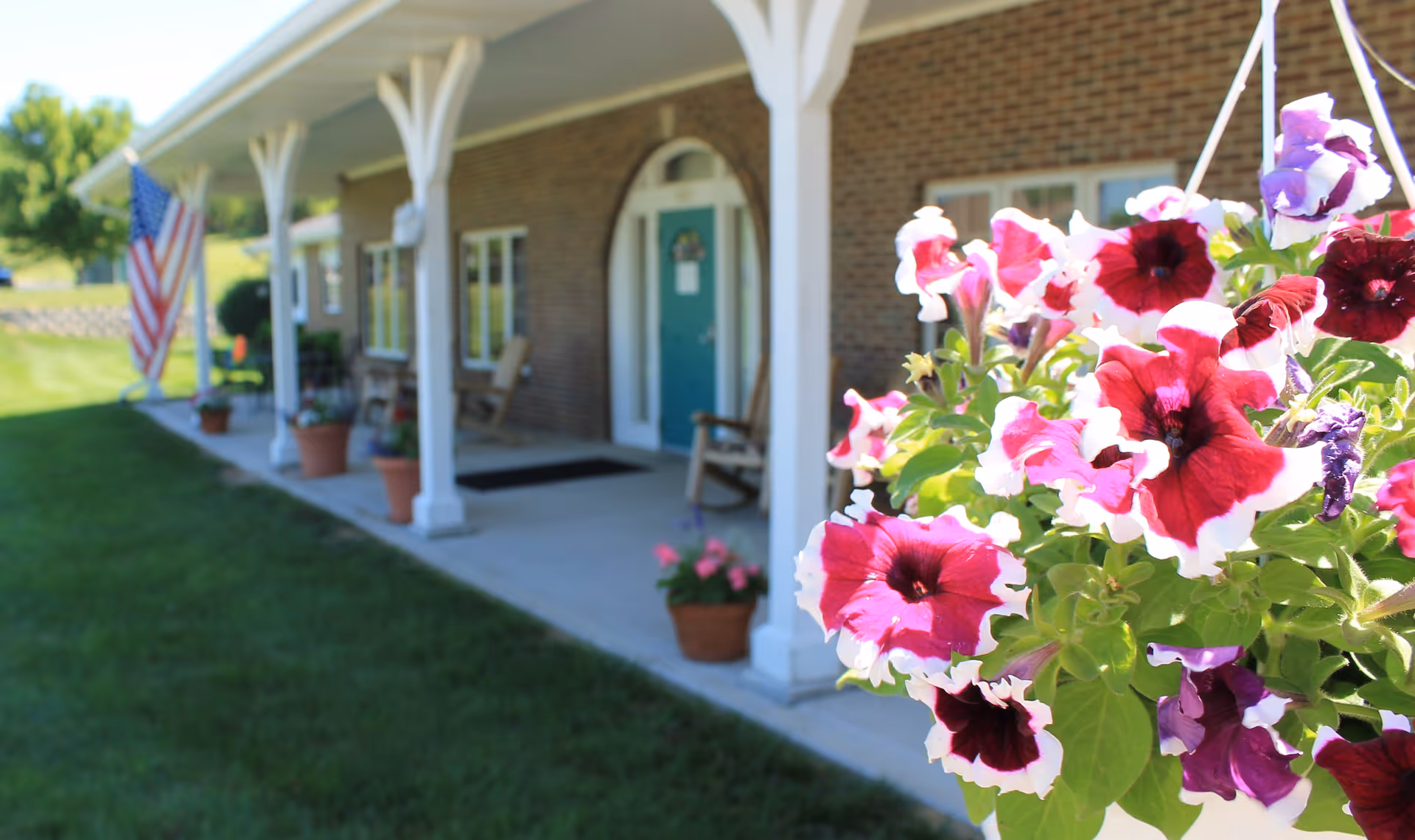 Close-up of vibrant pink and purple petunias in a hanging basket with a brick building porch in the background, featuring white columns, rocking chairs, potted plants, and an American flag.