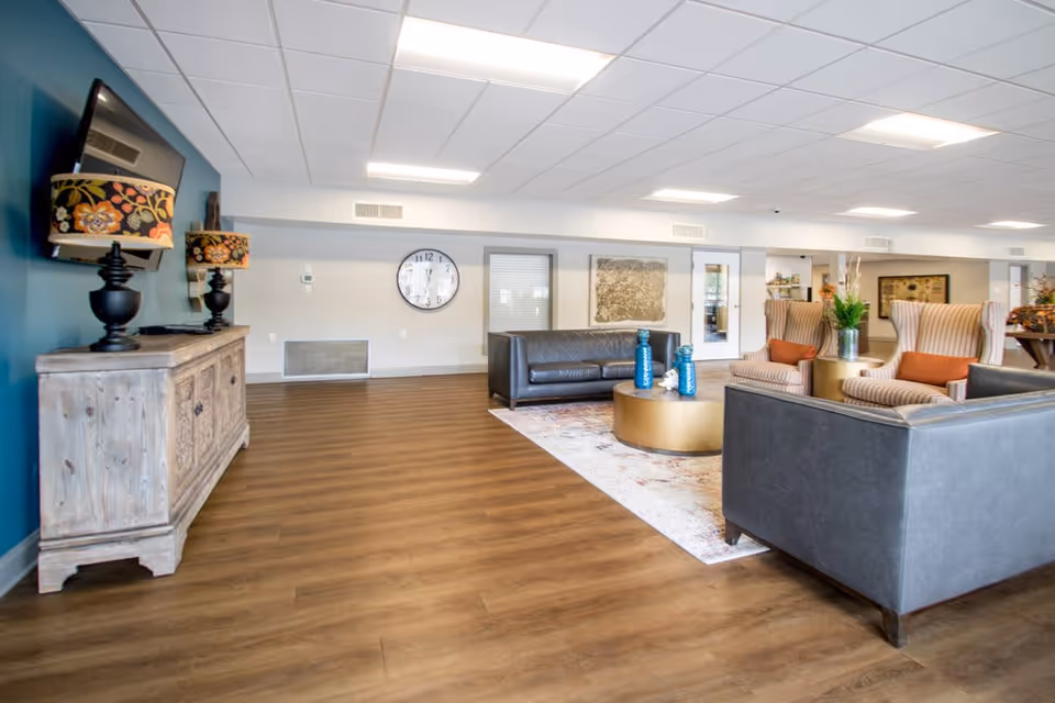 A spacious and well-lit living room area with wooden flooring, featuring two gray leather sofas, two striped armchairs with orange cushions, a round gold coffee table with decorative blue bottles, a large wall clock, a flat-screen TV mounted on a teal accent wall, and a wooden cabinet with two floral-patterned lamps.