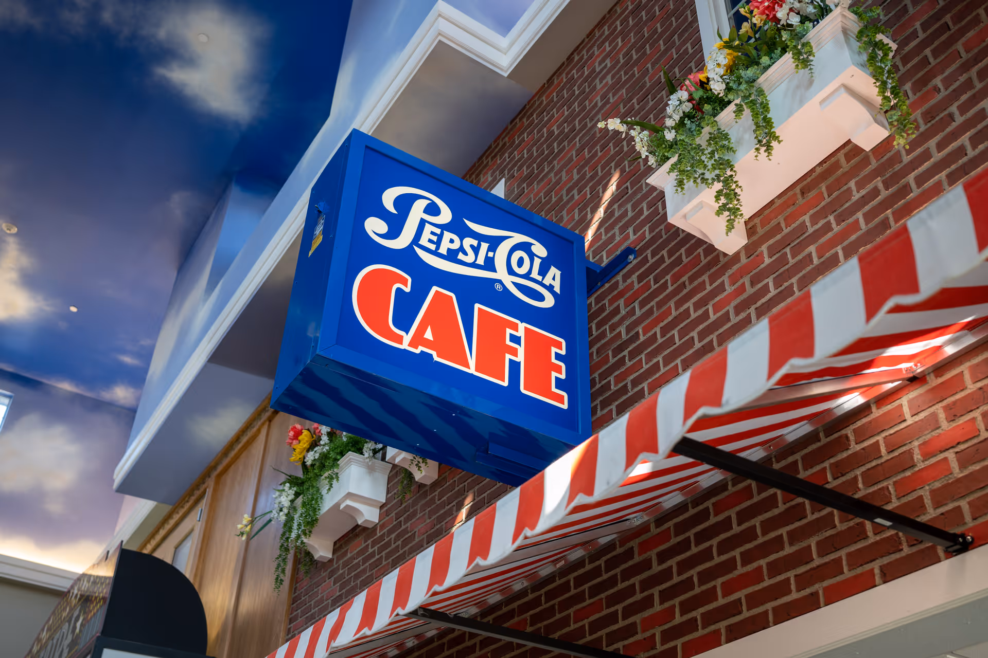 A blue and red Pepsi-Cola Cafe sign hanging on a brick wall above a red and white striped awning, with flower boxes containing green plants and flowers mounted on the wall. The ceiling above is painted with a sky and clouds motif.