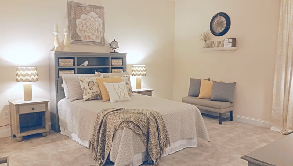 A cozy bedroom with a neatly made bed featuring multiple decorative pillows, including one with the word 'LOVE'. The bed has a textured beige bedspread and a woven throw blanket draped over the foot. On either side of the bed are wooden nightstands with matching lamps that have chevron-patterned shades. Above the bed is a large floral wall art. To the right, there is a small cushioned bench with three pillows in blue and yellow. A wall shelf above the bench holds a clock, a decorative 'HOPE' sign, and a small framed picture. The room has beige walls and carpet, with a curtain partially visible on the right side.