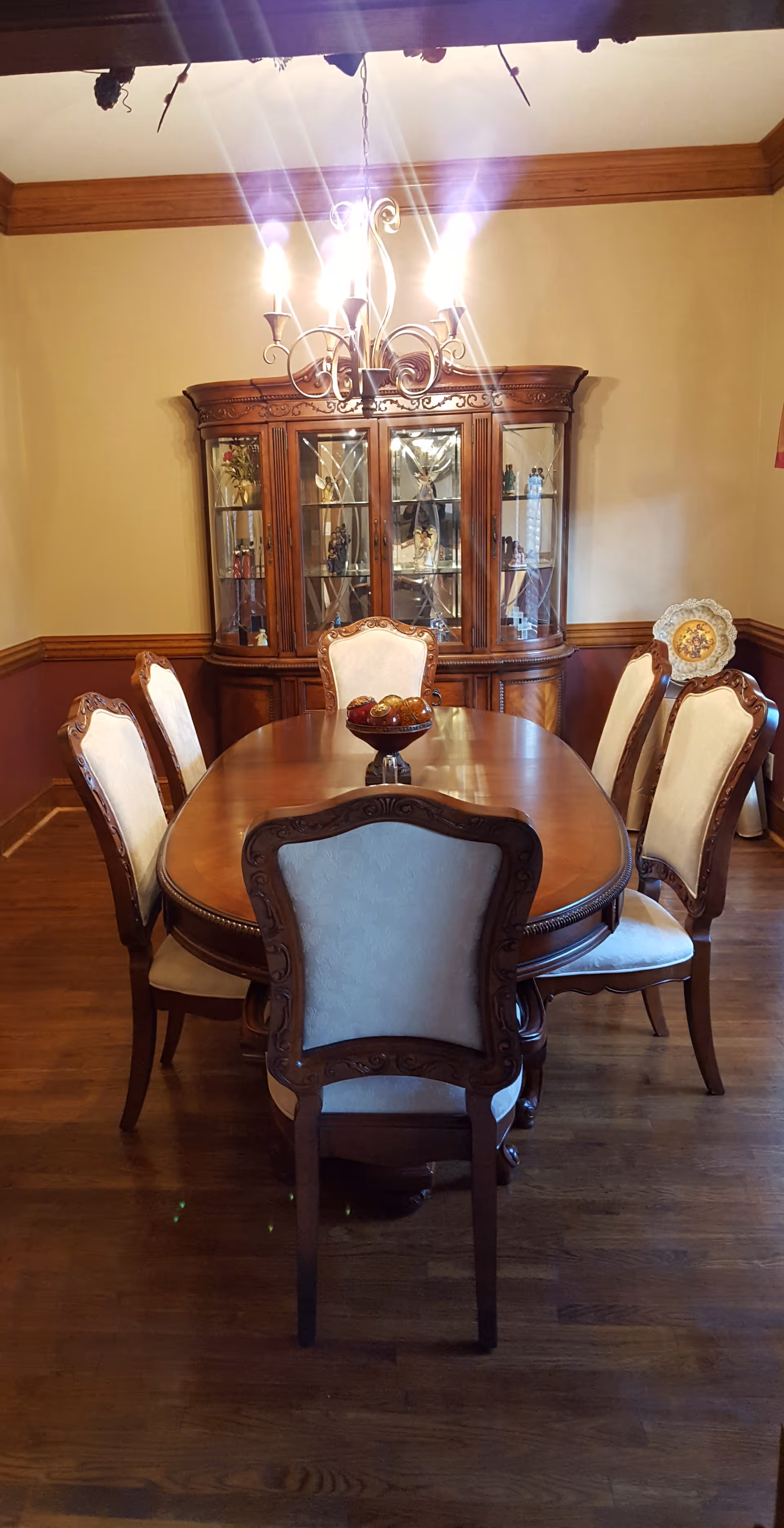 A formal dining room with a wooden dining table surrounded by six upholstered chairs. A decorative bowl with fruit is placed in the center of the table. Behind the table is a wooden china cabinet with glass doors displaying various decorative items. A chandelier with lit bulbs hangs above the table, and the room has wood trim and hardwood flooring.