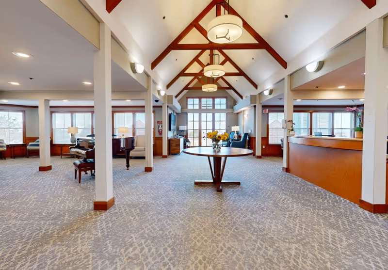 Spacious senior living facility common area with high vaulted ceiling featuring exposed wooden beams and hanging light fixtures. The room has large windows letting in natural light, a round wooden table with a vase of sunflowers in the center, a grand piano on the left, and a reception desk on the right. Comfortable seating areas and lamps are visible near the windows.
