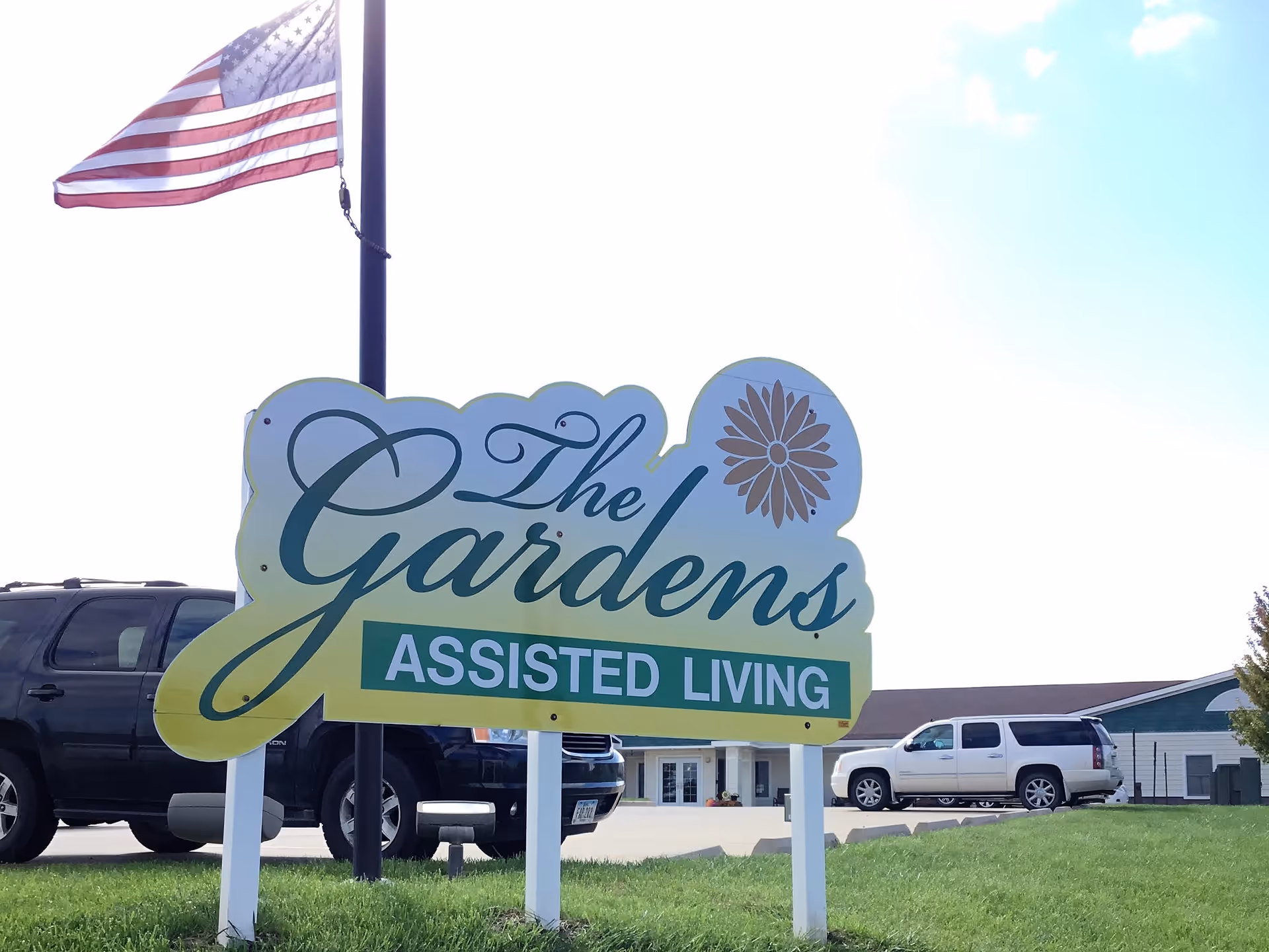 Outdoor view of a sign for The Gardens Assisted Living with an American flag on a pole behind it and parked cars in the background under a partly cloudy sky.