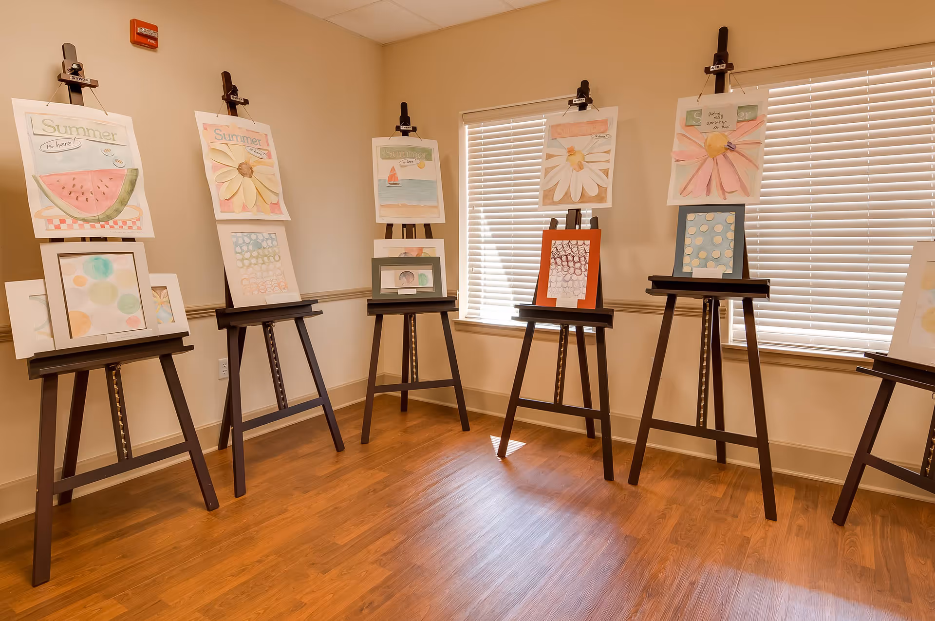A room with wooden floors and beige walls featuring several easels displaying colorful paintings of flowers, a watermelon slice, and abstract patterns. The room has two windows with white blinds partially closed, allowing natural light to enter.