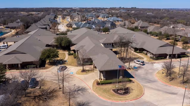 Aerial view of Keller Oaks Healthcare Center showing multiple single-story buildings with gray roofs arranged around curved driveways and parking areas, surrounded by leafless trees and residential houses in the background under a clear sky.