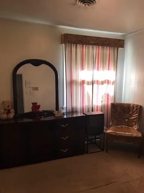 A room with a dark wooden dresser featuring a mirror, a teddy bear, and a red vase on top. Next to the dresser is a small black TV on a stand and a chair covered in plastic. A window with striped curtains is behind the chair.
