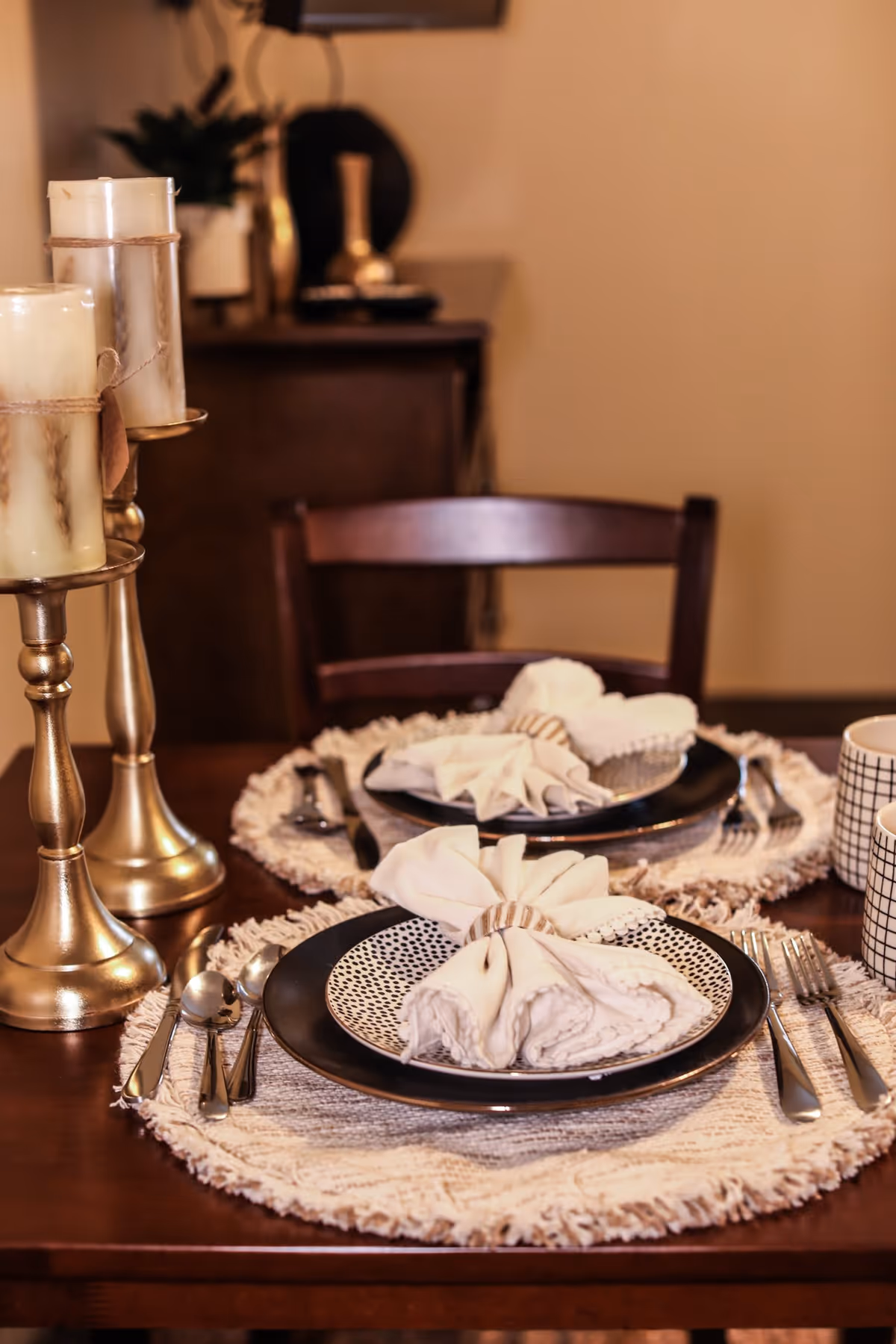 A close-up view of a dining table set for two with decorative plates, folded napkins, silverware, and two patterned mugs. Two tall gold candle holders with white candles are placed on the left side of the table. A wooden chair and a sideboard with decorative items are visible in the background.