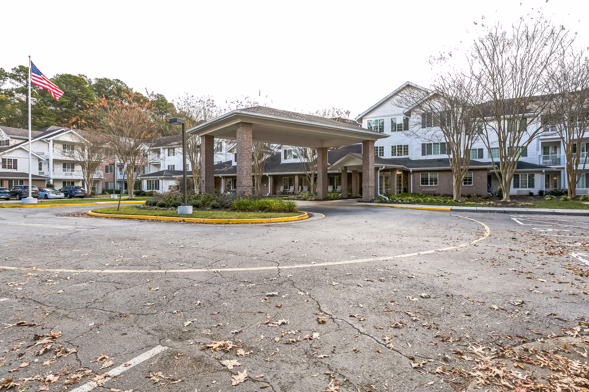 Front entrance of a multi-story senior living facility with a covered porte-cochere, parking circle, flagpole and landscaped grounds.