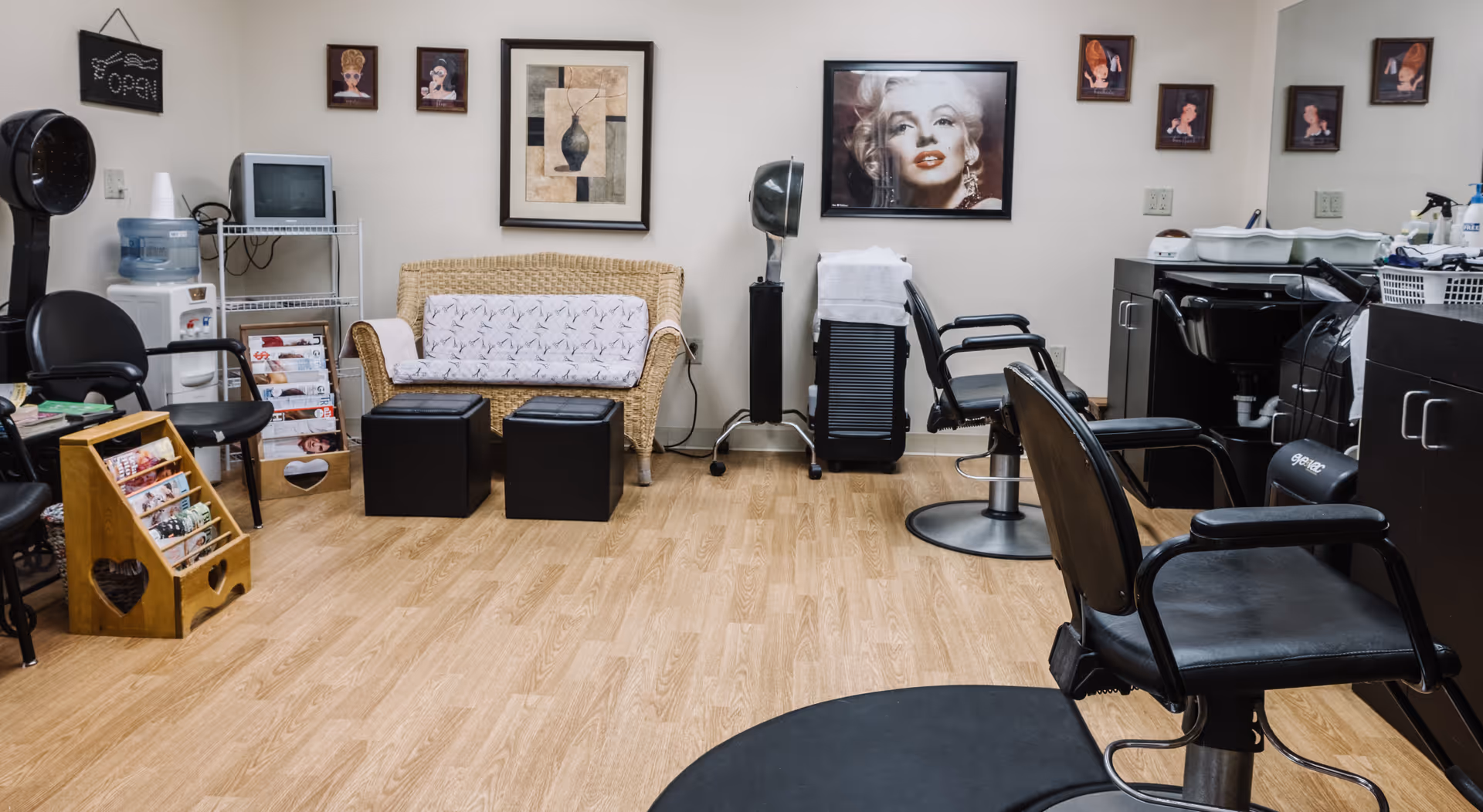 Interior of a senior living facility's salon area with black salon chairs, a hair dryer, a wicker loveseat with a patterned cover, two black ottomans, a water cooler, a small TV on a metal rack, and framed pictures on the walls including a large portrait of Marilyn Monroe.