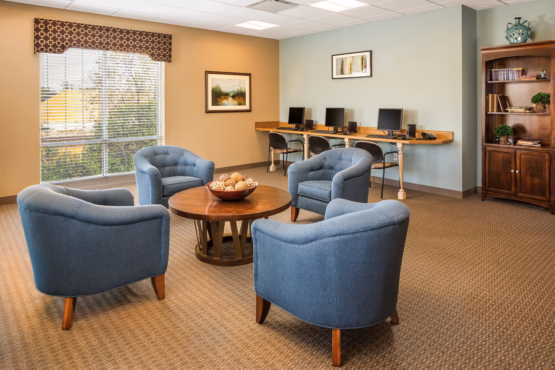 A cozy common area with four blue upholstered armchairs arranged around a round wooden coffee table with decorative balls. Behind the chairs, there is a long wooden desk with three desktop computers and chairs. The room has beige walls, a large window with blinds and a patterned valance, two framed paintings on the walls, and a wooden bookshelf with books and plants.