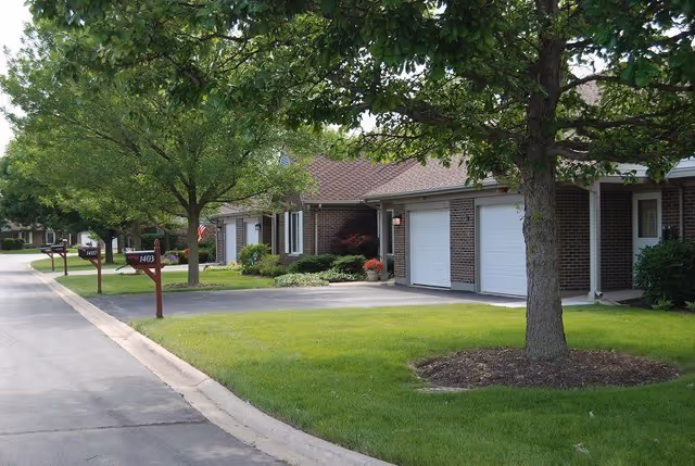 A quiet residential street in a senior living community with single-story brick homes, each with a garage and mailbox. The street is lined with green lawns and trees providing shade.