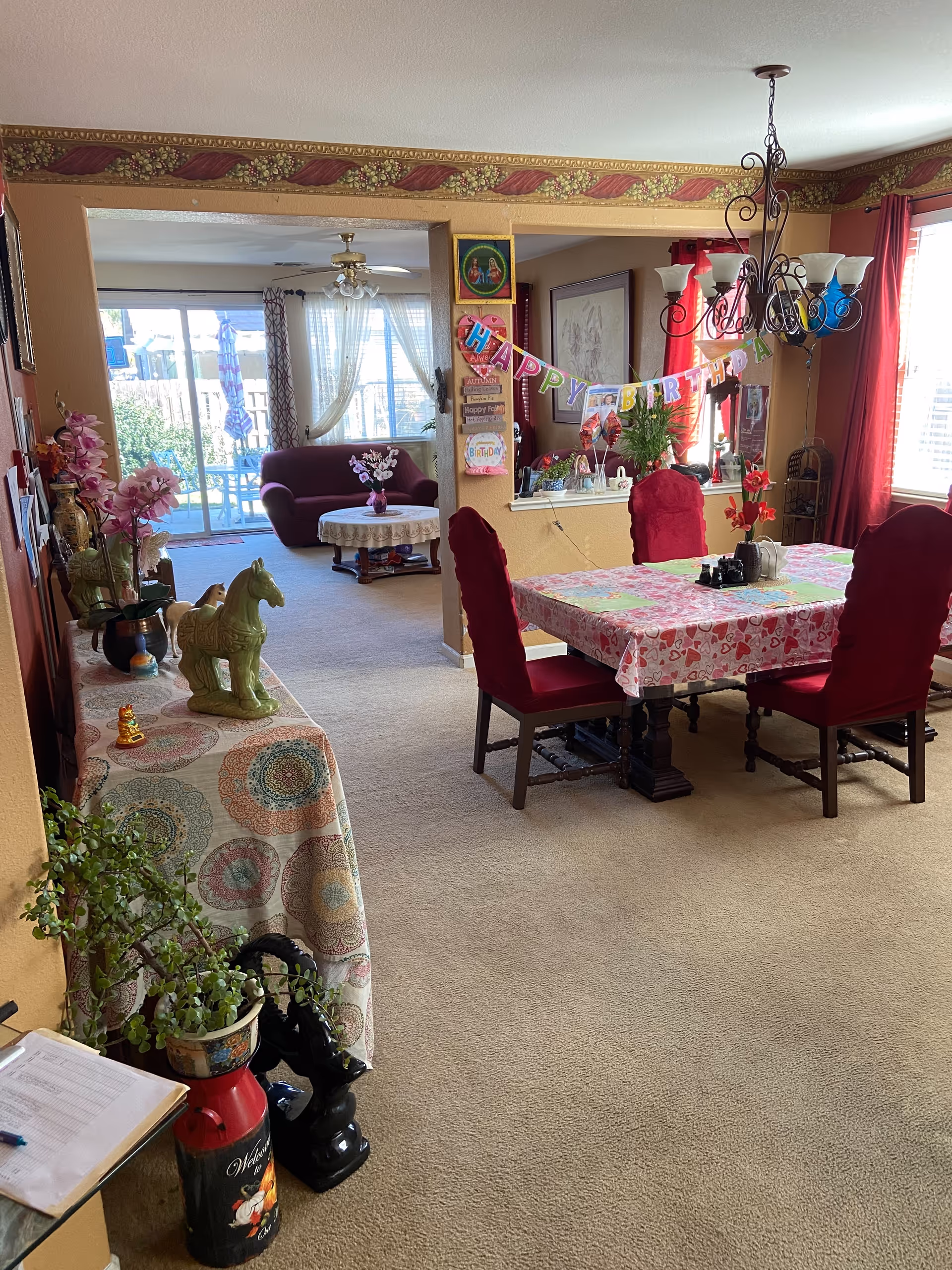 Interior view of a senior living facility showing a dining area with a table covered in a pink and red heart-patterned tablecloth and four red upholstered chairs. A chandelier hangs above the table. In the background, there is a living room area with a maroon sofa and a round table with a lace tablecloth and a flower vase. The room is decorated with a 'Happy Birthday' banner, plants, and various decorative items. Large windows with curtains allow natural light to fill the space.