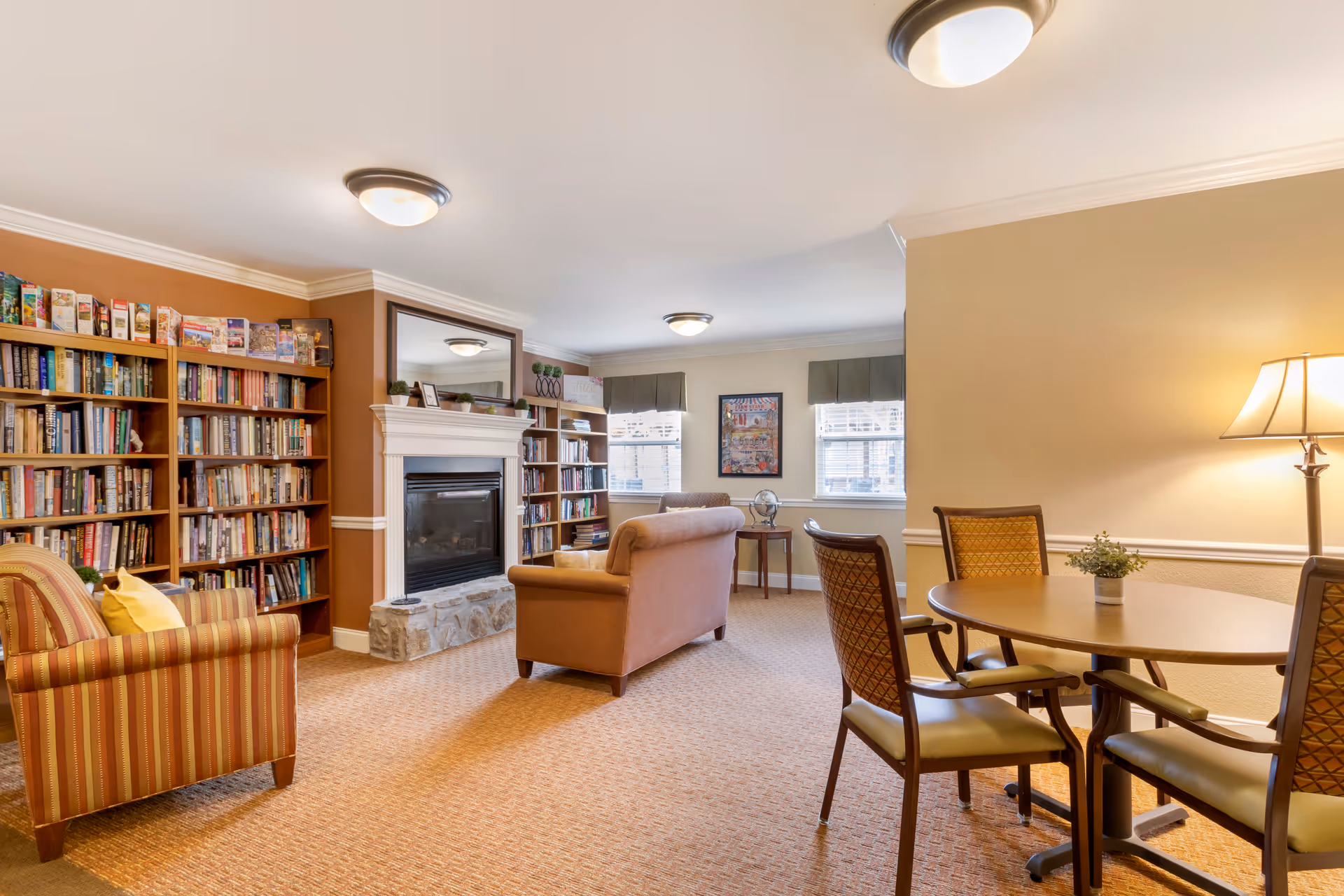 A cozy living room area in a senior living facility featuring a striped armchair with a yellow pillow, a brown sofa, a fireplace with a white mantel and stone base, bookshelves filled with books, a round table with four chairs, a floor lamp, and two windows with blinds and valances.