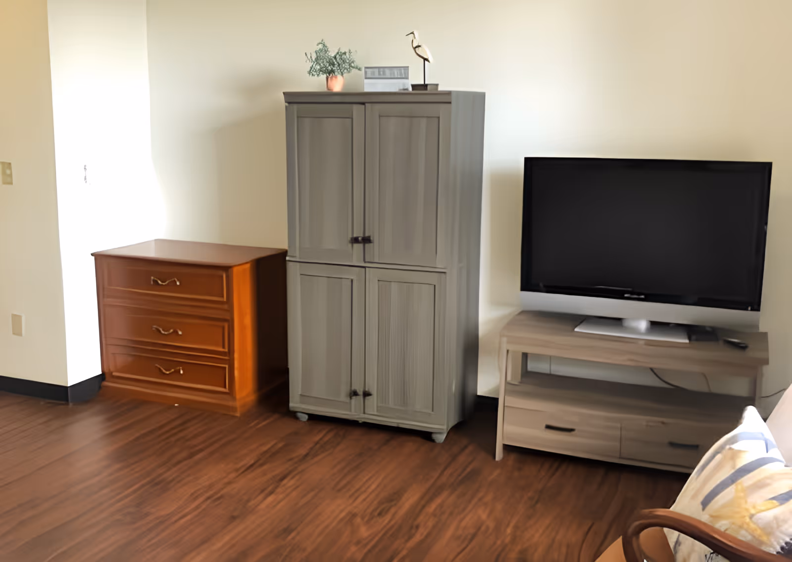 A corner of a room with wooden flooring featuring a small wooden dresser with three drawers, a gray cabinet with two sets of double doors, a flat-screen TV on a light wood TV stand with two drawers, and part of a chair with a cushion in the foreground.