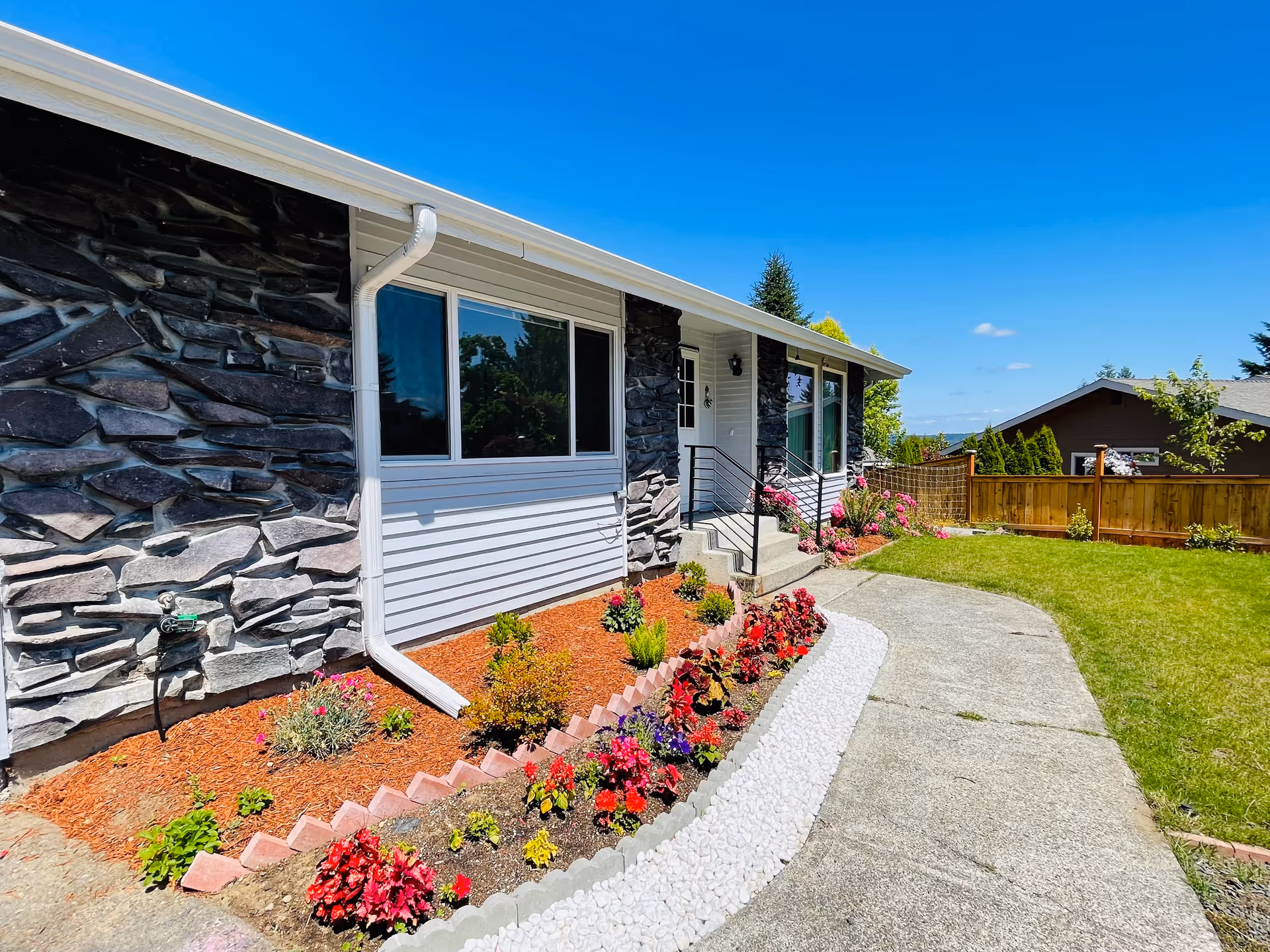 Exterior view of a single-story building with stone and white siding walls, large windows, a small set of stairs leading to a door, and a well-maintained garden with colorful flowers and shrubs along a curved concrete pathway under a clear blue sky.
