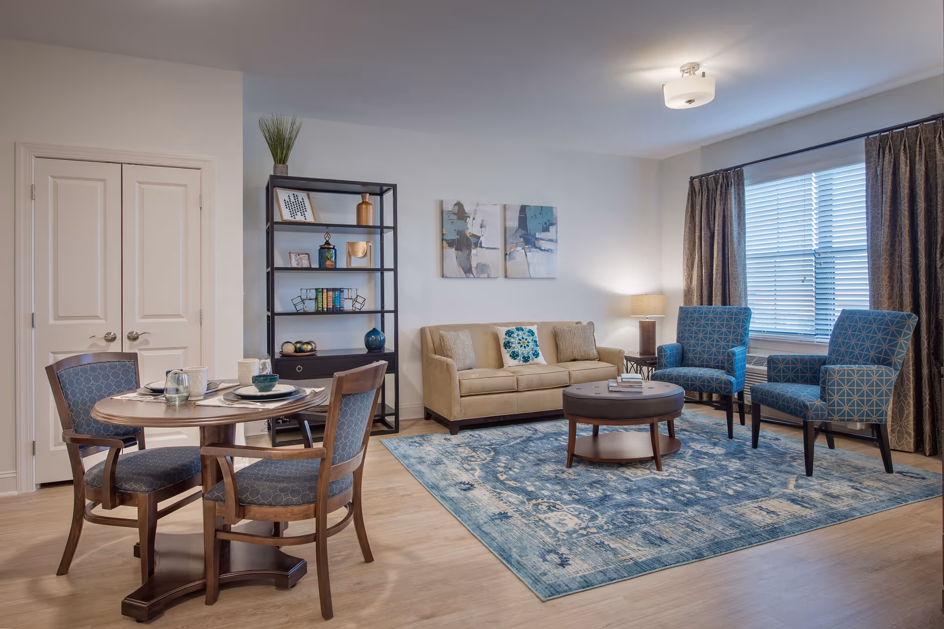 Furnished living room and dining area with a beige sofa, two blue patterned armchairs, round coffee table, dining table with four chairs, bookshelf, and large window.