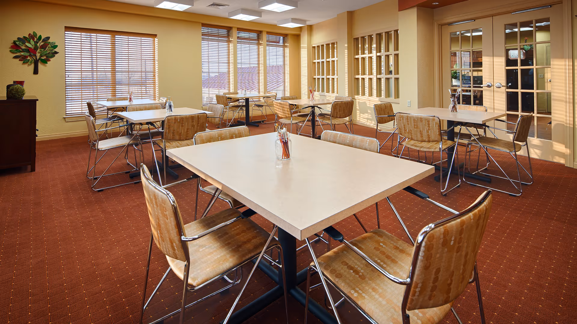 A dining room with several square tables and chairs arranged neatly on a red carpeted floor. The room has large windows with wooden blinds allowing natural light to enter. There is a decorative wall piece shaped like a tree and a cabinet on the left side. The walls are painted a light yellow color, and there are glass-paned double doors on the right side.