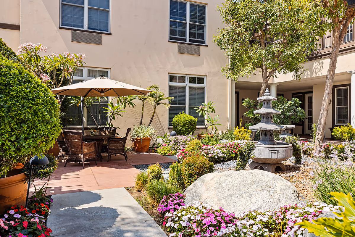 Outdoor courtyard area at a senior living facility with a stone pagoda-style fountain, colorful flower beds, potted plants, trees, and a patio seating area with a table, chairs, and a large umbrella. The building exterior with windows is visible in the background.