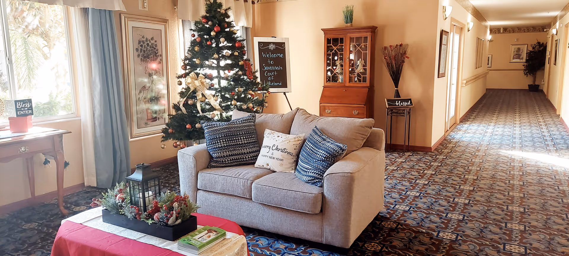 A cozy seating area in a hallway decorated for Christmas with a small Christmas tree adorned with ornaments and a gold ribbon. A beige loveseat with three decorative pillows sits in front of the tree. To the left, there is a small wooden table with a potted plant and a sign that says 'Bless Enter'. A red table with a festive centerpiece and books is in the foreground. The hallway has patterned carpet and cream-colored walls with framed artwork and a wooden cabinet. A chalkboard sign near the tree reads 'Welcome to Savannah Court of Lakeland'.