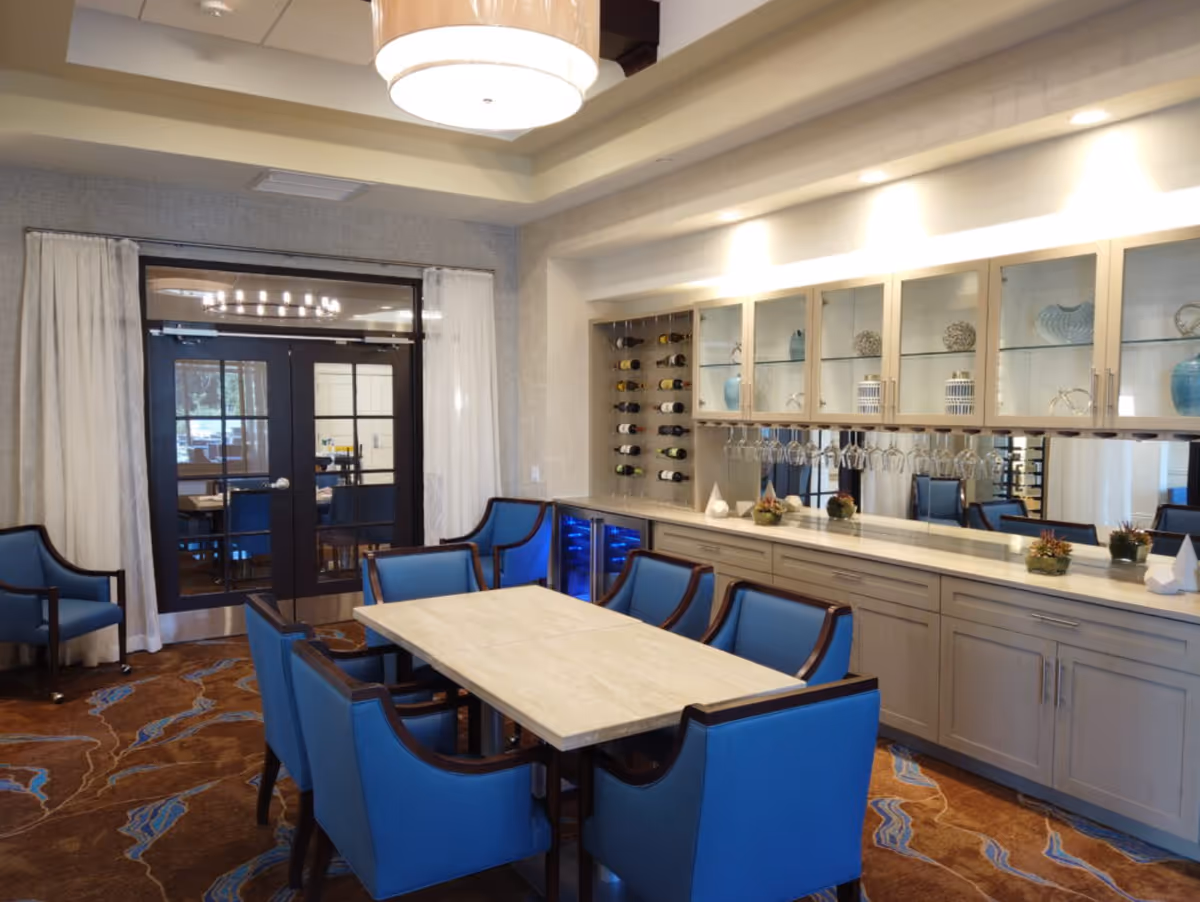A dining room with a rectangular table surrounded by eight blue upholstered chairs. Behind the table is a built-in cabinet with glass doors displaying decorative items and a wine rack with bottles. Wine glasses hang upside down under the cabinet. The room has a patterned carpet and double glass doors with white curtains on either side.