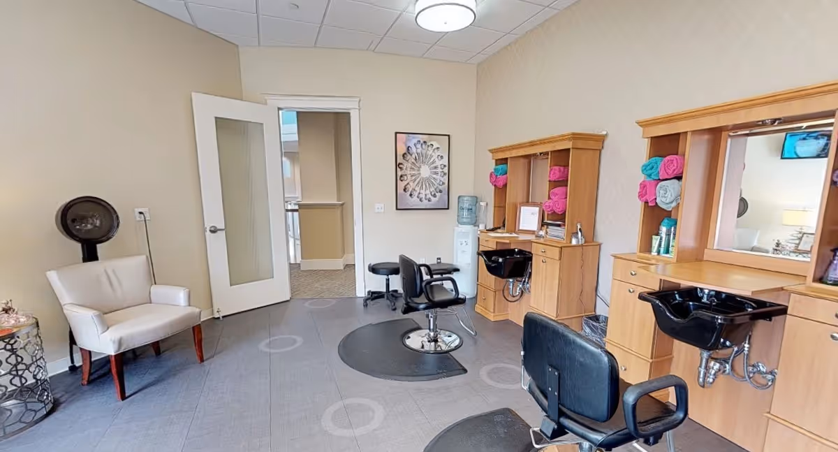 Interior view of a salon area in a senior living facility with two black salon chairs in front of wooden stations equipped with sinks and mirrors. Colorful rolled towels are placed on shelves, and a white armchair with a hair dryer is positioned near a small round table. A water cooler and framed artwork are visible on the beige walls.