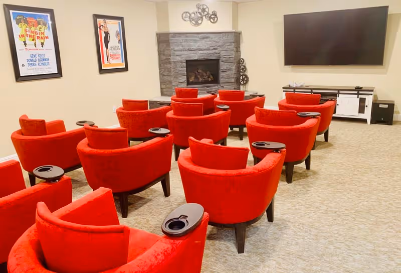 A cozy media room with multiple red upholstered chairs arranged in rows facing a large flat-screen TV mounted on the wall. The room features a stone fireplace beneath a decorative metal wall art piece. Two framed vintage movie posters hang on the left wall.