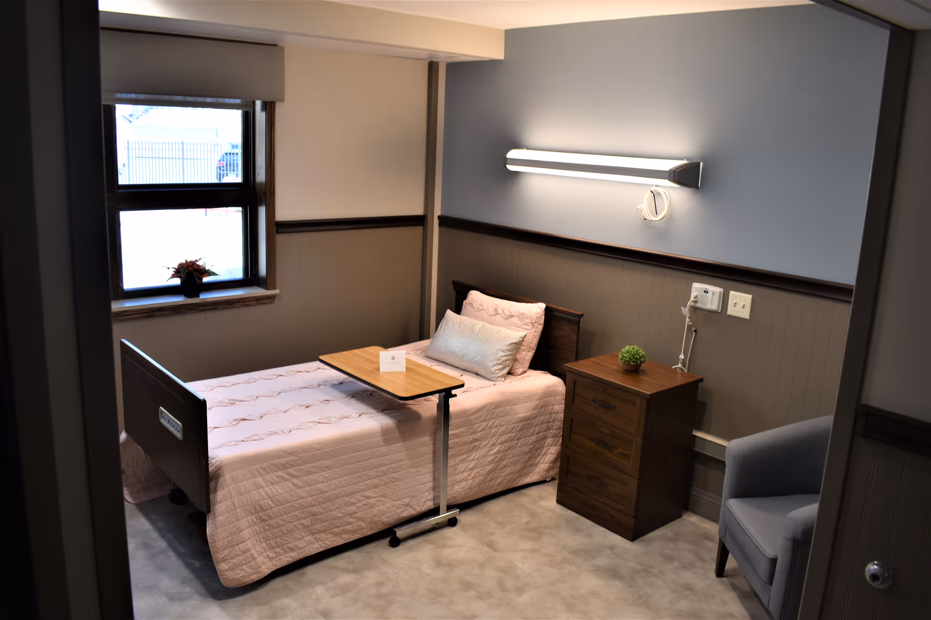 A small, neatly arranged bedroom in a medical care facility featuring a single bed with a pink quilt and pillows, a wooden bedside table with a small green plant, a gray armchair, a window with a flower pot on the sill, and a wall-mounted light above the bed.