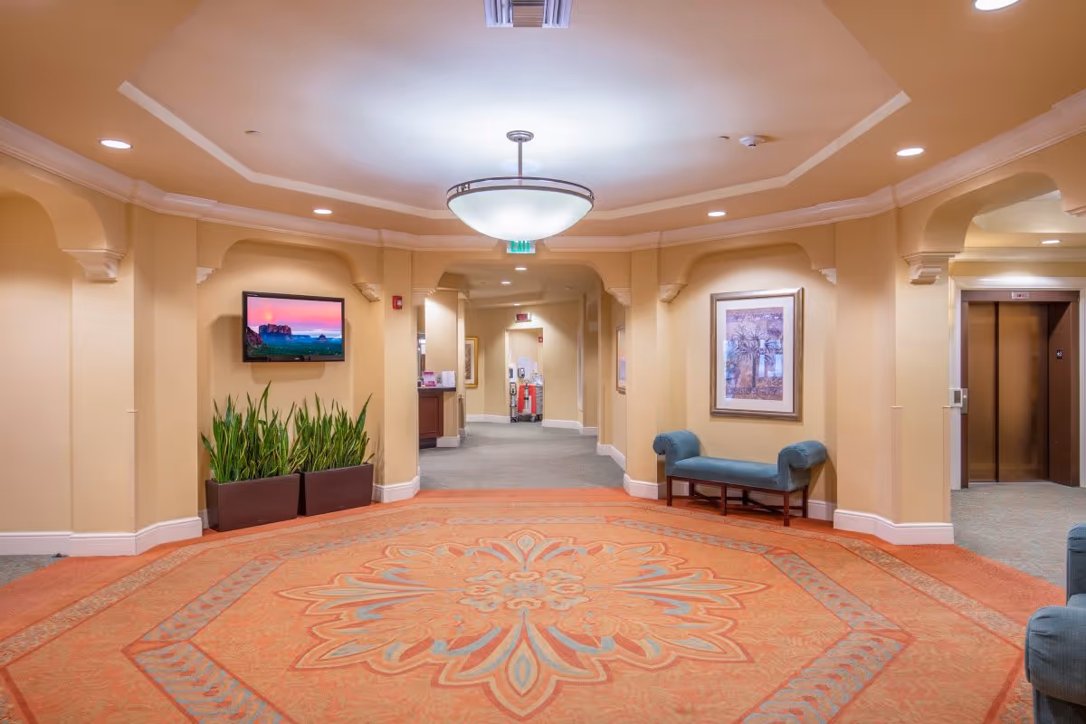 A spacious, well-lit hallway in a senior living facility with beige walls and an ornate orange and blue patterned carpet. The ceiling features a large circular light fixture. There is a flat-screen TV mounted on the left wall above two rectangular planters with green plants. On the right wall, there is a framed artwork above a blue upholstered bench. An elevator with closed doors is visible on the far right, and the hallway extends further into the building.