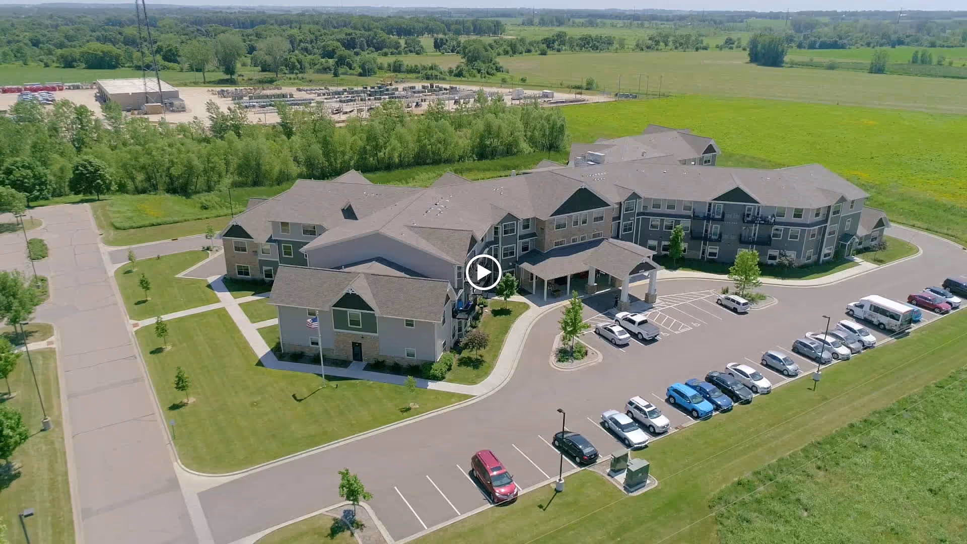 Aerial view of Legacy of Farmington Senior Living facility showing a large, multi-wing building surrounded by green lawns and trees. There is a parking lot with several cars parked in front of the building. The surrounding area includes open fields and some industrial structures in the distance.