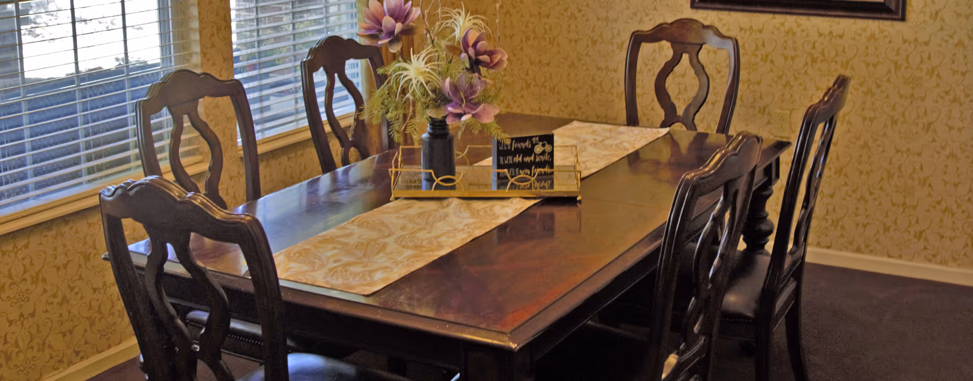 A dining room with a dark wooden rectangular table surrounded by six matching chairs. The table has a beige patterned runner and a centerpiece with purple flowers and greenery in a black vase. The room has patterned wallpaper and large windows with blinds.