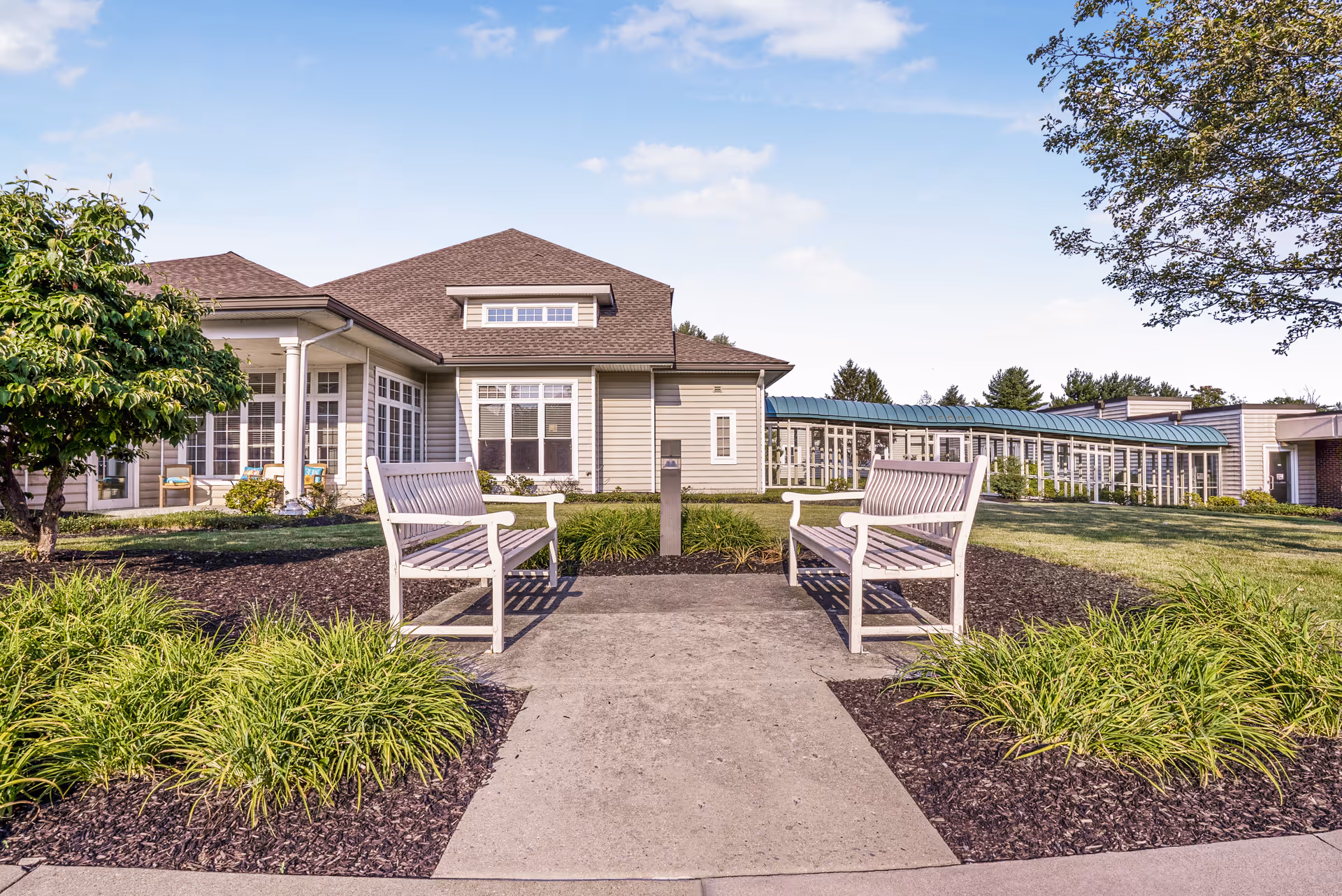 Two white benches facing each other on a walkway in front of a single-story senior living building with landscaped grounds.