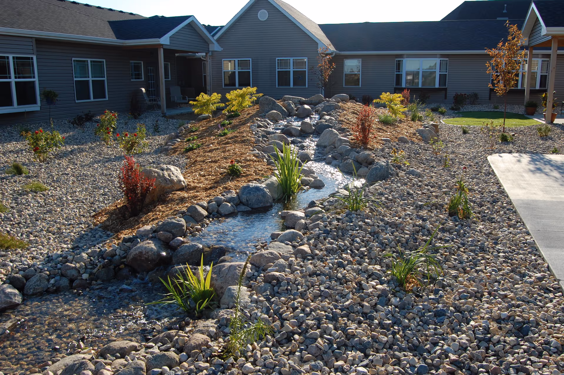 Outdoor garden area at StoneyBrook Suites Assisted Living featuring a small stream with rocks and plants, surrounded by gravel and landscaping, with the facility building in the background.