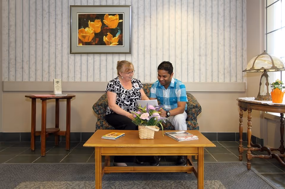 A woman and a man sitting together on a floral patterned couch in a cozy room. They are looking at a tablet and smiling. In front of them is a wooden coffee table with a basket of flowers and some magazines. The room has patterned wallpaper, a framed picture of yellow tulips on the wall, a wooden side table with a lamp, and a window letting in natural light.