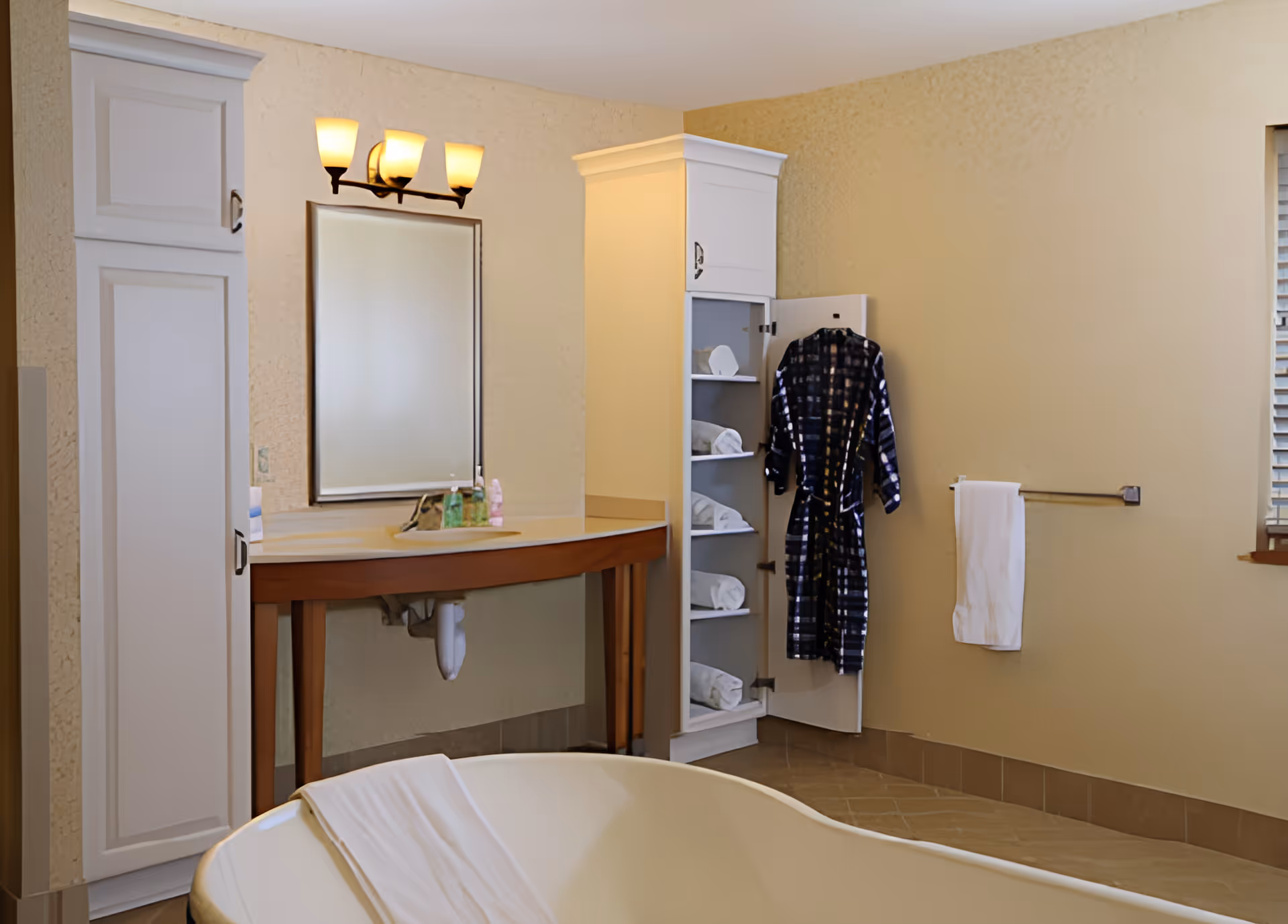A bathroom interior featuring a white bathtub with a towel draped over the edge, a wooden vanity with a sink and mirror above it, a tall white cabinet with shelves holding rolled towels, and a black and white checkered robe hanging on the cabinet door. A towel rack with a white towel is mounted on the beige wall next to a window with blinds.
