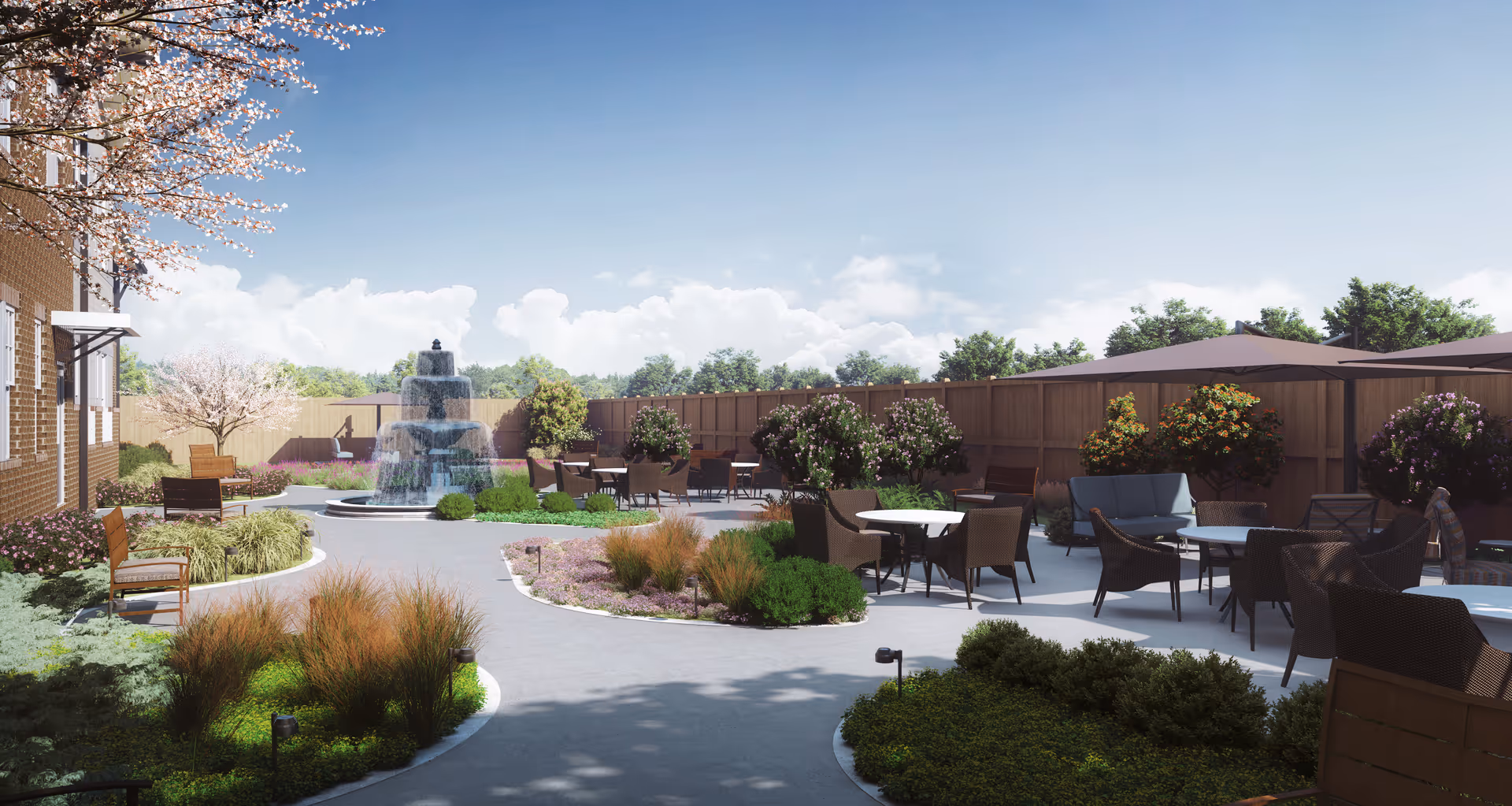 Outdoor patio area with multiple seating arrangements including tables and chairs, surrounded by various plants and bushes. A multi-tiered water fountain is visible in the background near a wooden fence. The sky is clear with some clouds, and there are flowering trees and shrubs around the patio.