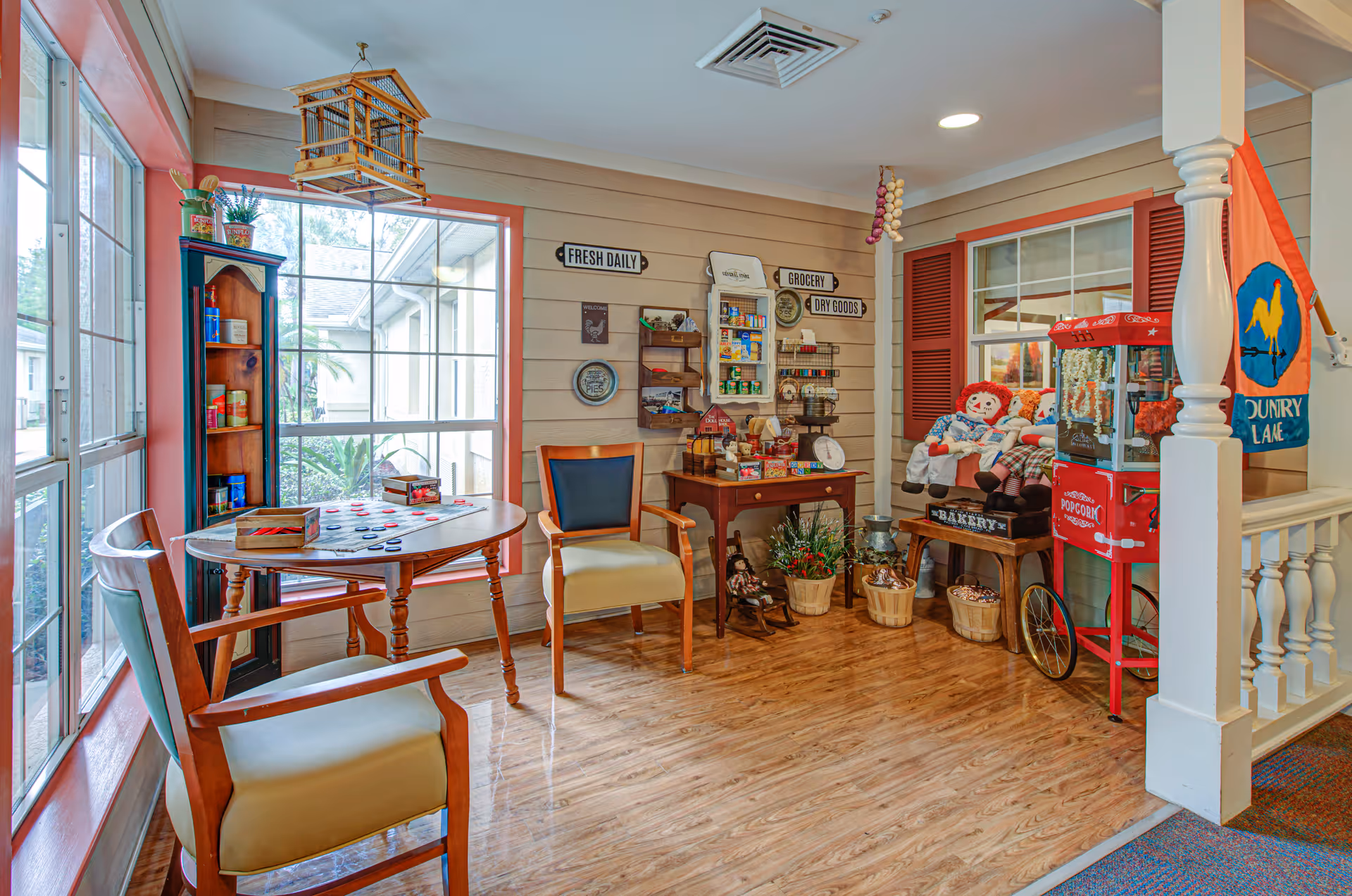 A cozy, well-lit corner of a senior living facility featuring a small wooden table with two chairs, a tall shelving unit with canned goods, and a wall decorated with signs reading 'Fresh Daily,' 'Grocery,' and 'Dry Goods.' There is a small table with various items, baskets with plants, and a popcorn machine next to a window with red shutters. Two Raggedy Ann dolls sit on a bench near the popcorn machine, and a colorful flag with a rooster and the words 'Country Lane' hangs on the wall.
