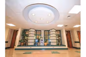 Interior view of a healthcare facility lobby or common area with a circular ceiling light fixture, potted plants, and a water fountain against a wall with large windows and green trim.