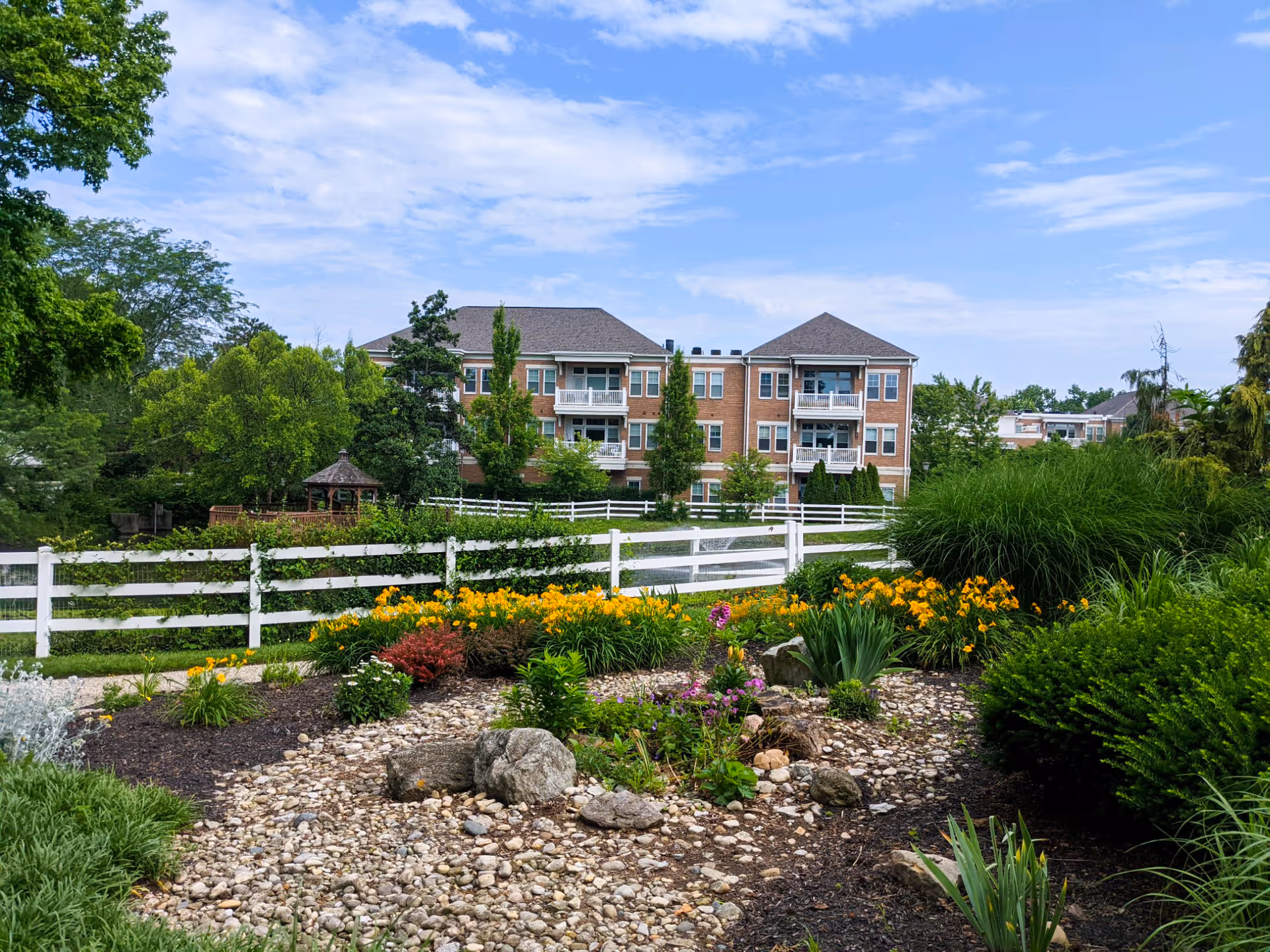 A landscaped garden area with colorful flowers, shrubs, and rocks in the foreground, a white wooden fence, and a multi-story brick building with balconies in the background under a partly cloudy blue sky.