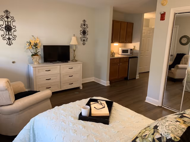 A cozy senior living room area with a beige armchair, a white dresser holding a TV, a lamp, and a flower vase. The room has wooden flooring and light-colored walls with decorative wall hangings. A small kitchenette with wooden cabinets, a microwave, and a mini fridge is visible in the background. A bed with a light blue quilt and a tray holding a book, glasses, and a cup is in the foreground. A mirrored closet door reflects part of the room.