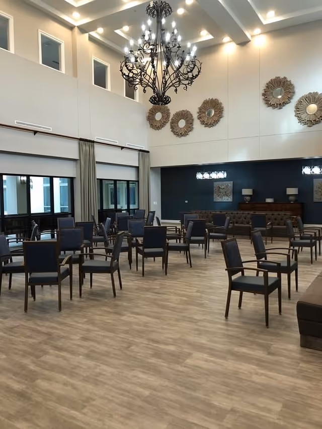 Spacious common area with multiple dark wooden chairs arranged in rows on a light wood floor. The room features high ceilings with a large ornate chandelier, decorative circular wall mirrors, and large windows with curtains allowing natural light.