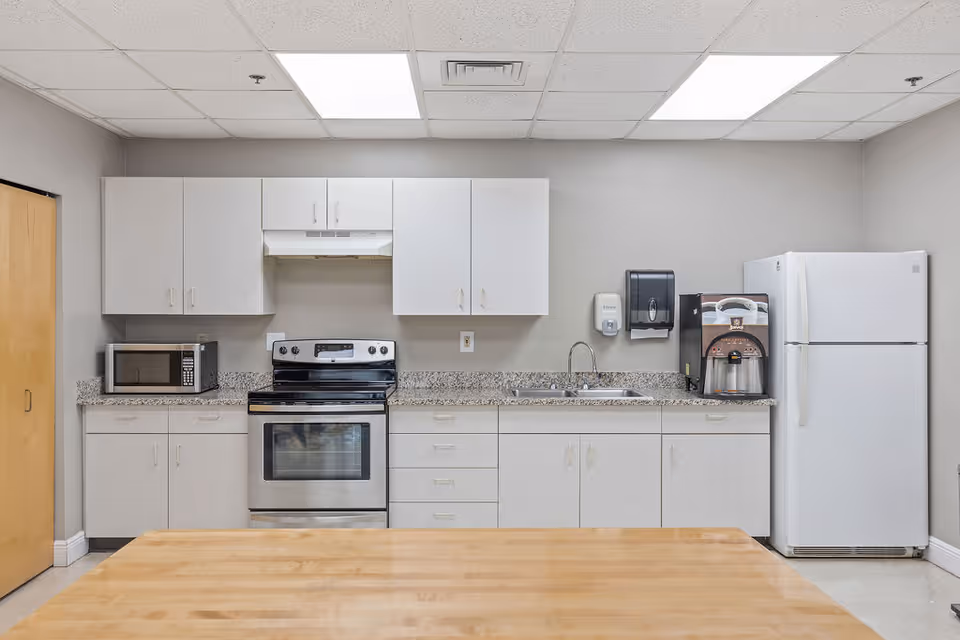 A clean and modern kitchen with white cabinets, a stainless steel electric stove and oven, a microwave on the left countertop, a double sink with a soap dispenser and paper towel holder on the wall above it, a coffee machine on the right countertop, and a white refrigerator. The ceiling has recessed lighting panels and the floor is light-colored.
