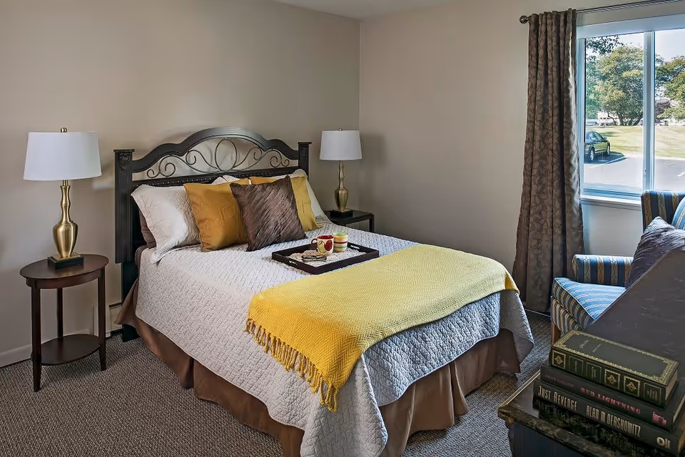 A cozy bedroom with a neatly made bed featuring a white quilt, brown bed skirt, and yellow throw blanket. There are multiple pillows in white, yellow, and brown colors. A tray with a cup and a small jar is placed on the bed. On either side of the bed are matching gold lamps on dark wooden nightstands. A window with brown curtains lets in natural light, and a striped armchair is positioned near the window. A stack of books is visible on a dark piece of furniture in the foreground.